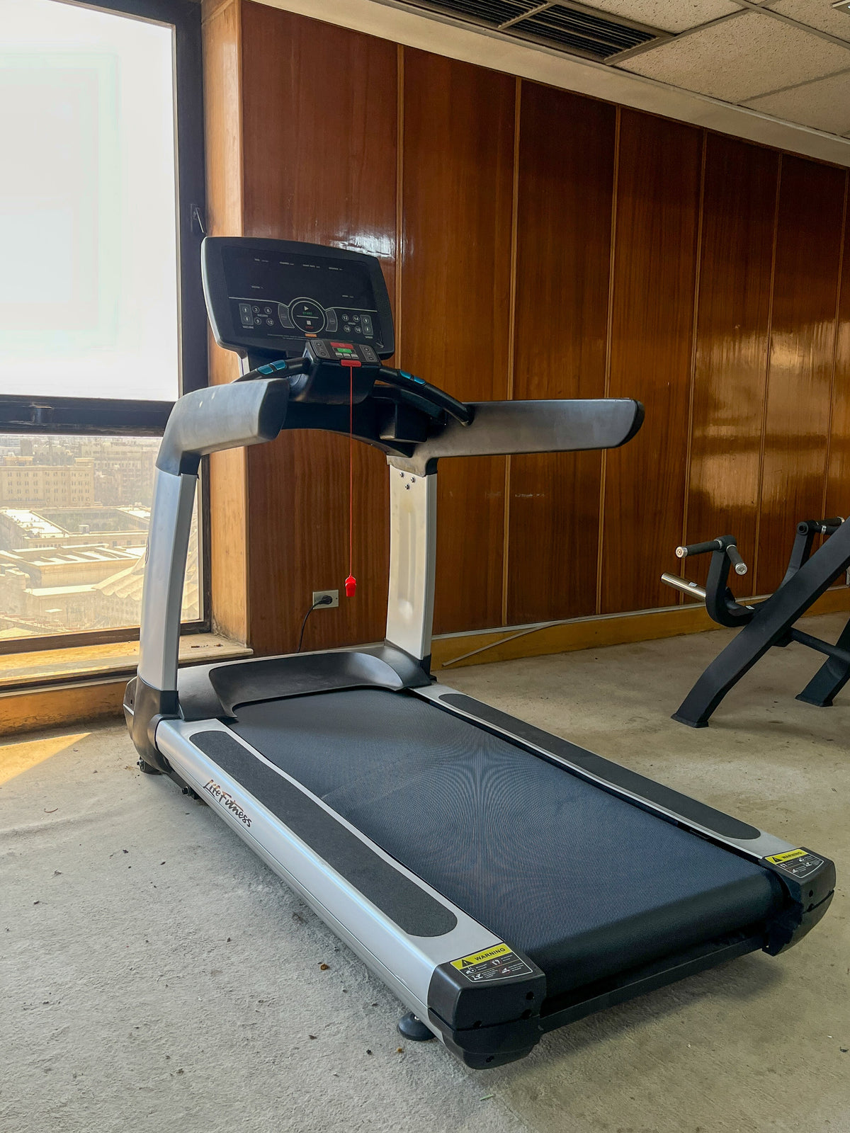 Lifefitness treadmill with a digital display console is shown in an indoor gym setting with other exercise equipment in the background.